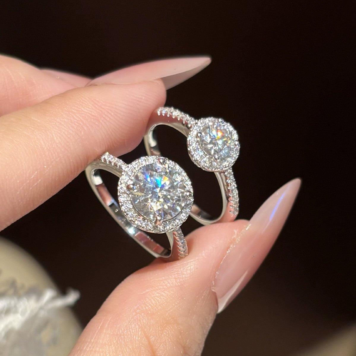 Two moissanite halo rings held between fingers, close-up of the pavé halo and shoulders