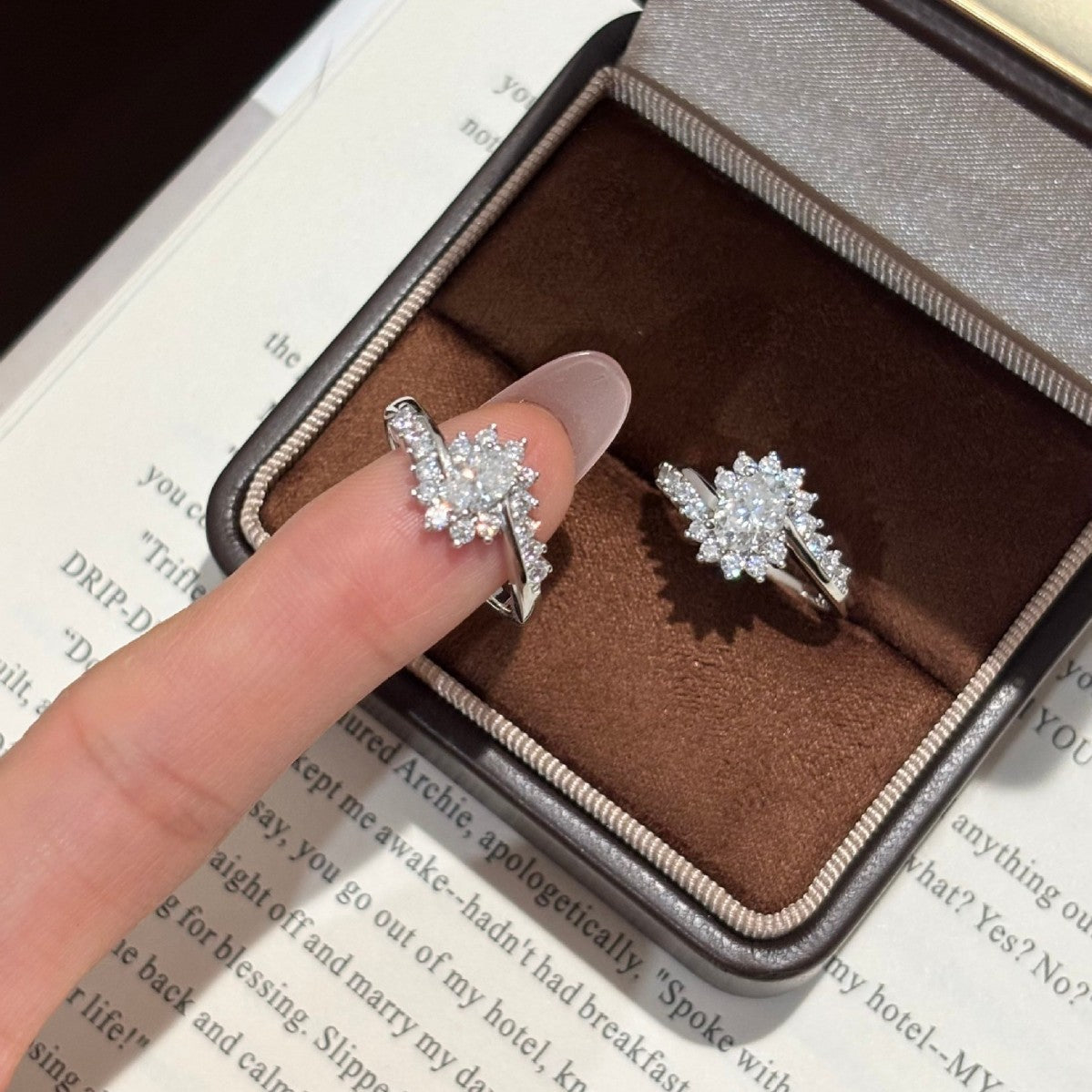 Sunburst moissanite ring held on a fingertip beside a second ring in a brown ring box